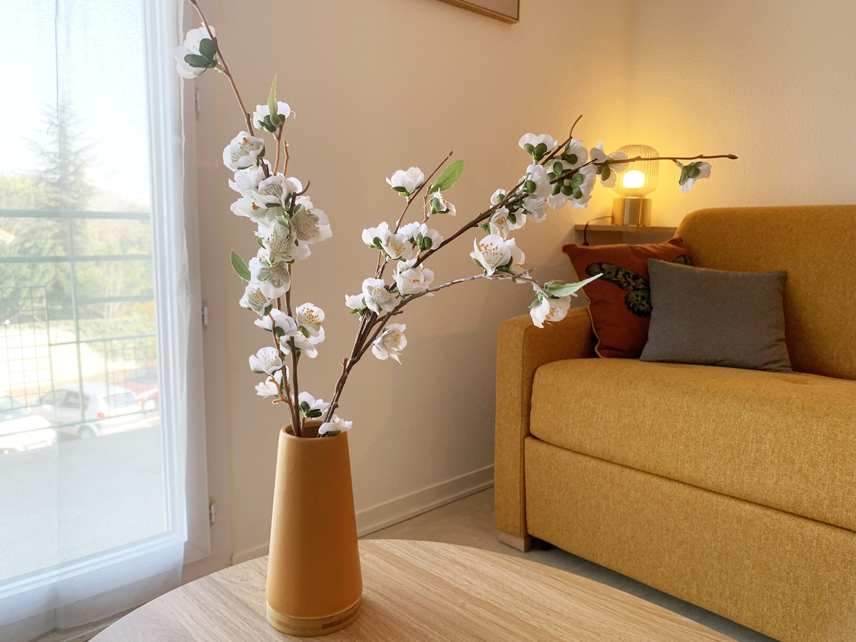 A cozy living area features a mustard-yellow couch adorned with cushions. A light-colored coffee table holds a vase with delicate white flowers. Soft natural light enters through the nearby window, enhancing the inviting atmosphere of the space.
