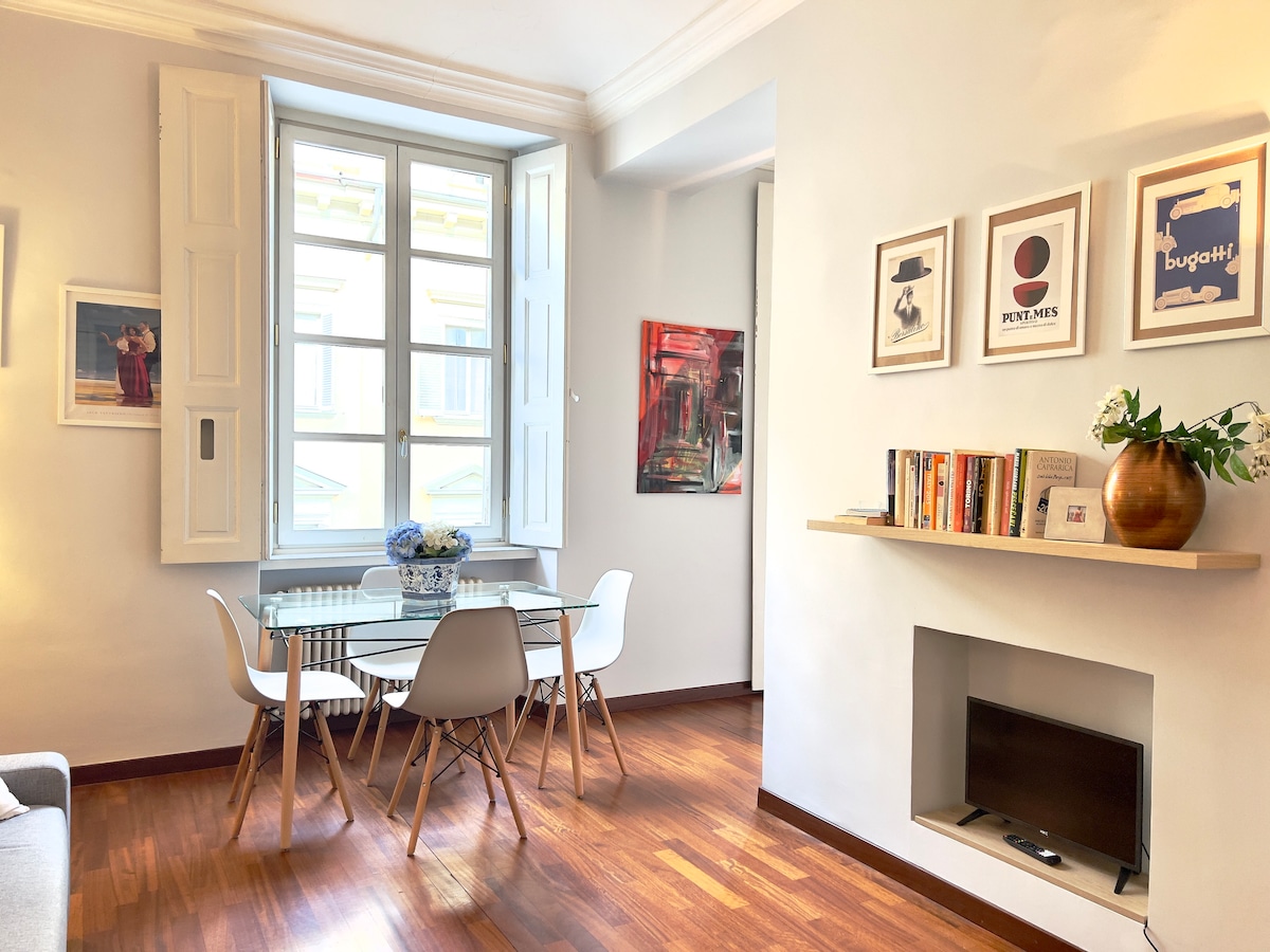 A well-lit dining area is shown, featuring a glass table surrounded by white chairs. Art pieces hang on the walls, complemented by wooden shelving holding books. Natural light filters through large windows with wooden shutters, highlighting the warm wooden floor.