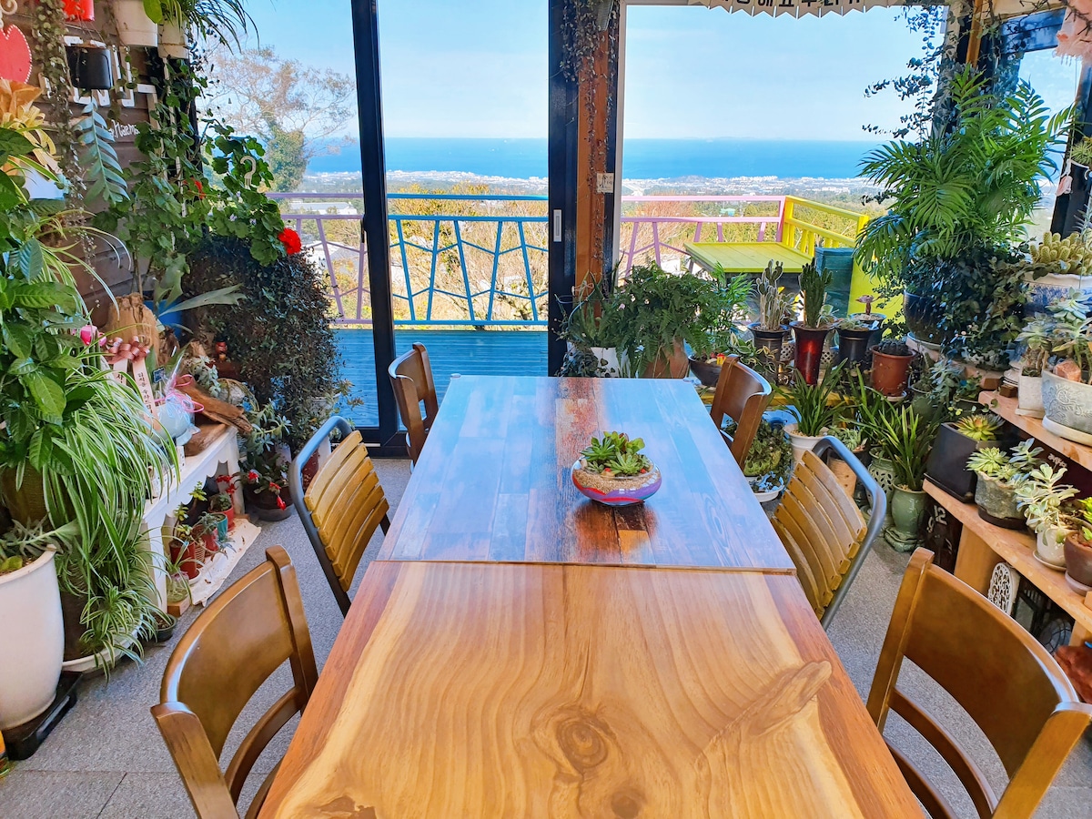A spacious dining area features a large wooden table surrounded by several chairs. Lush greenery and potted plants decorate the space, providing a vibrant backdrop. Expansive glass doors lead to a stunning view of the ocean, enhancing the inviting atmosphere.