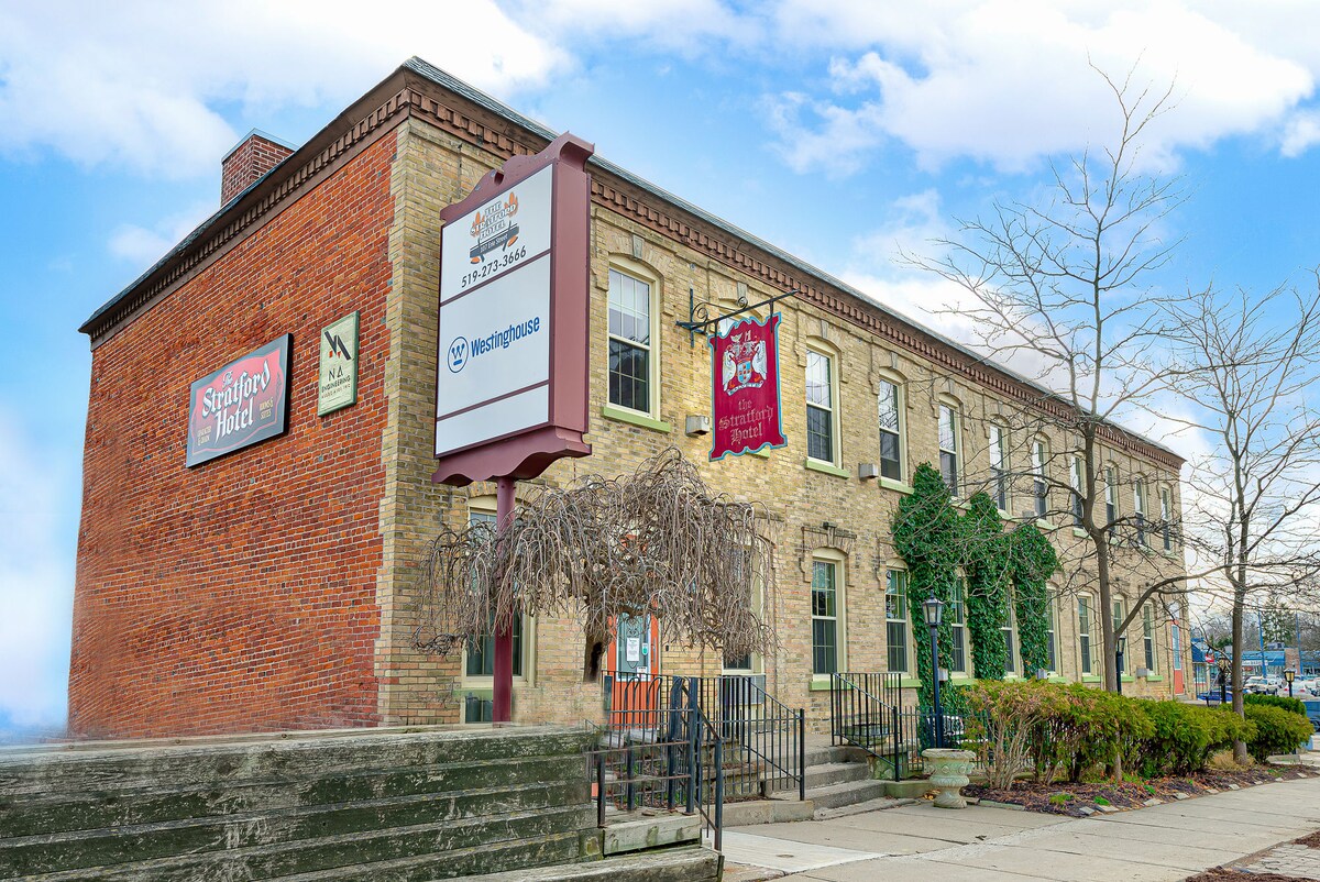 The exterior of The Stratford Hotel is depicted, showcasing a brick facade and large windows framed by greenery. Signage is visible, indicating the hotel name. The welcoming entrance is highlighted by accessible steps leading up to the door, with potted plants flanking the entrance.