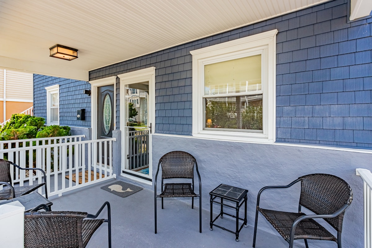 A covered front patio is featured, equipped with four black wicker chairs surrounding a small square table. The entrance is marked by a decorative door with a round window, and light landscaping can be seen in the background.