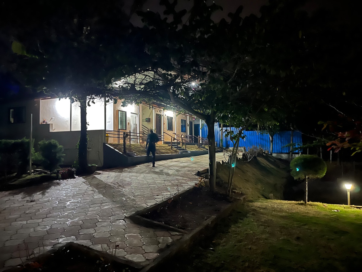 The villa is softly illuminated at night, surrounded by trees and pathways. Warm light glows from the windows, highlighting the entrance. A pathway lined with stone leads to the front door, while discreet outdoor lighting highlights the landscape.