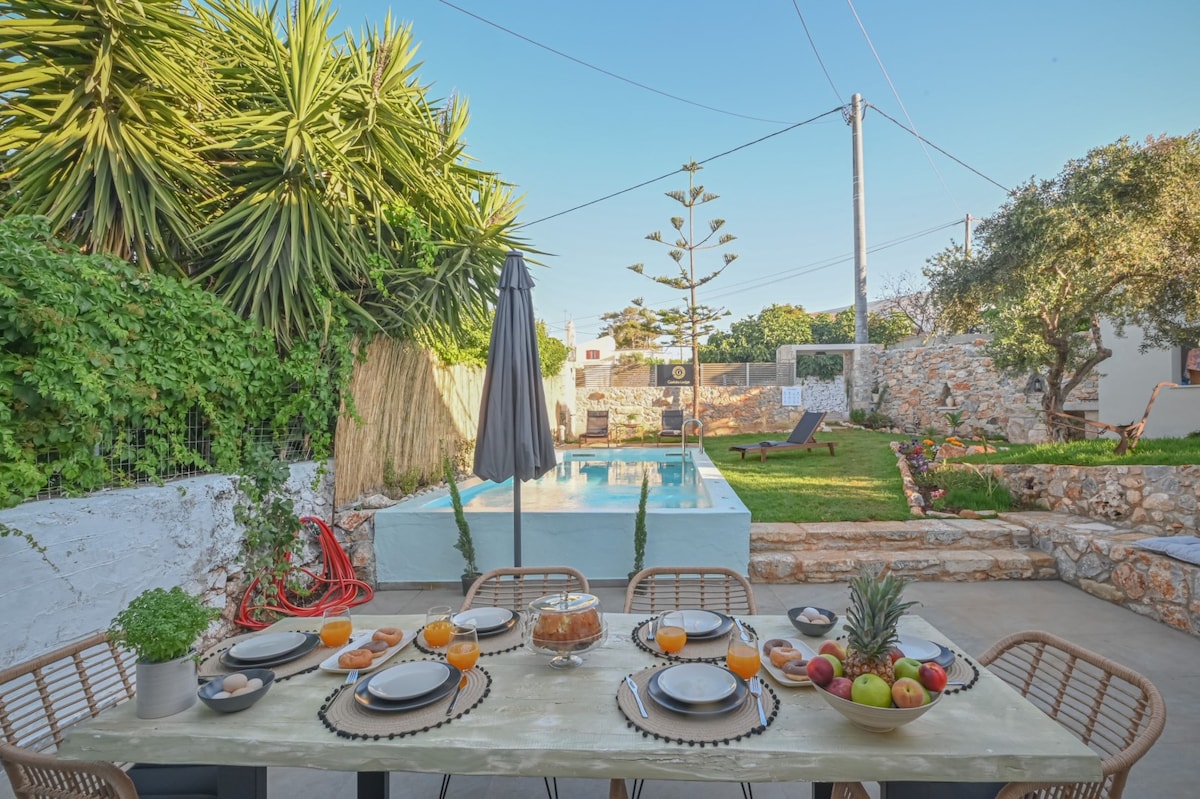 An outdoor dining area is presented with a large table set with plates, glasses, and fruit. A private pool is visible in the background, surrounded by lush greenery and garden plants. A sun umbrella and lounge chair are positioned nearby, creating a relaxed atmosphere.