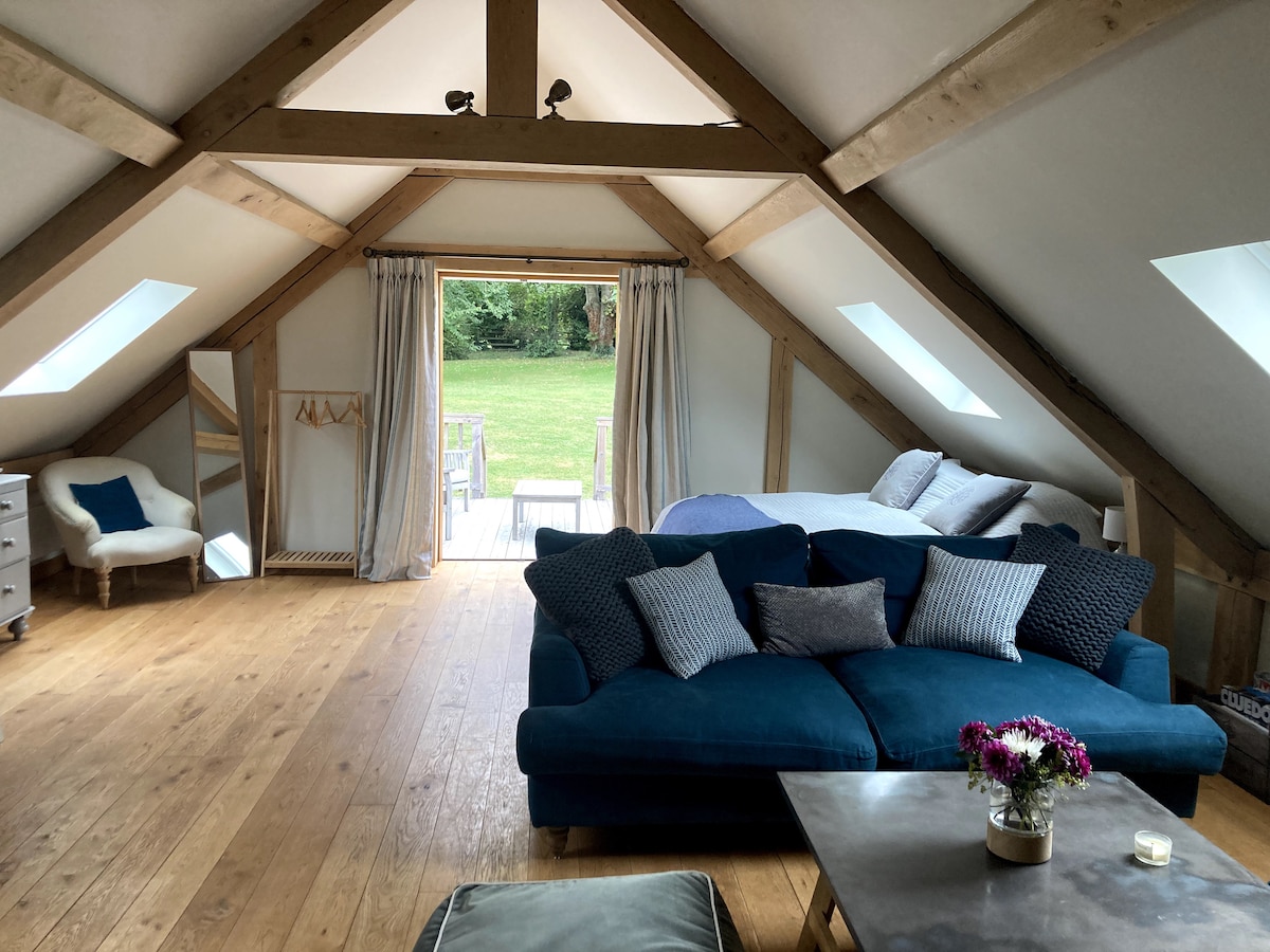 An open-plan living area features a plush blue sofa arranged around a wooden coffee table. Natural light streams in through vaulted ceilings and skylights, illuminating the space. A private terrace with outdoor seating can be seen through large glass doors, alongside a cozy bed in the background.