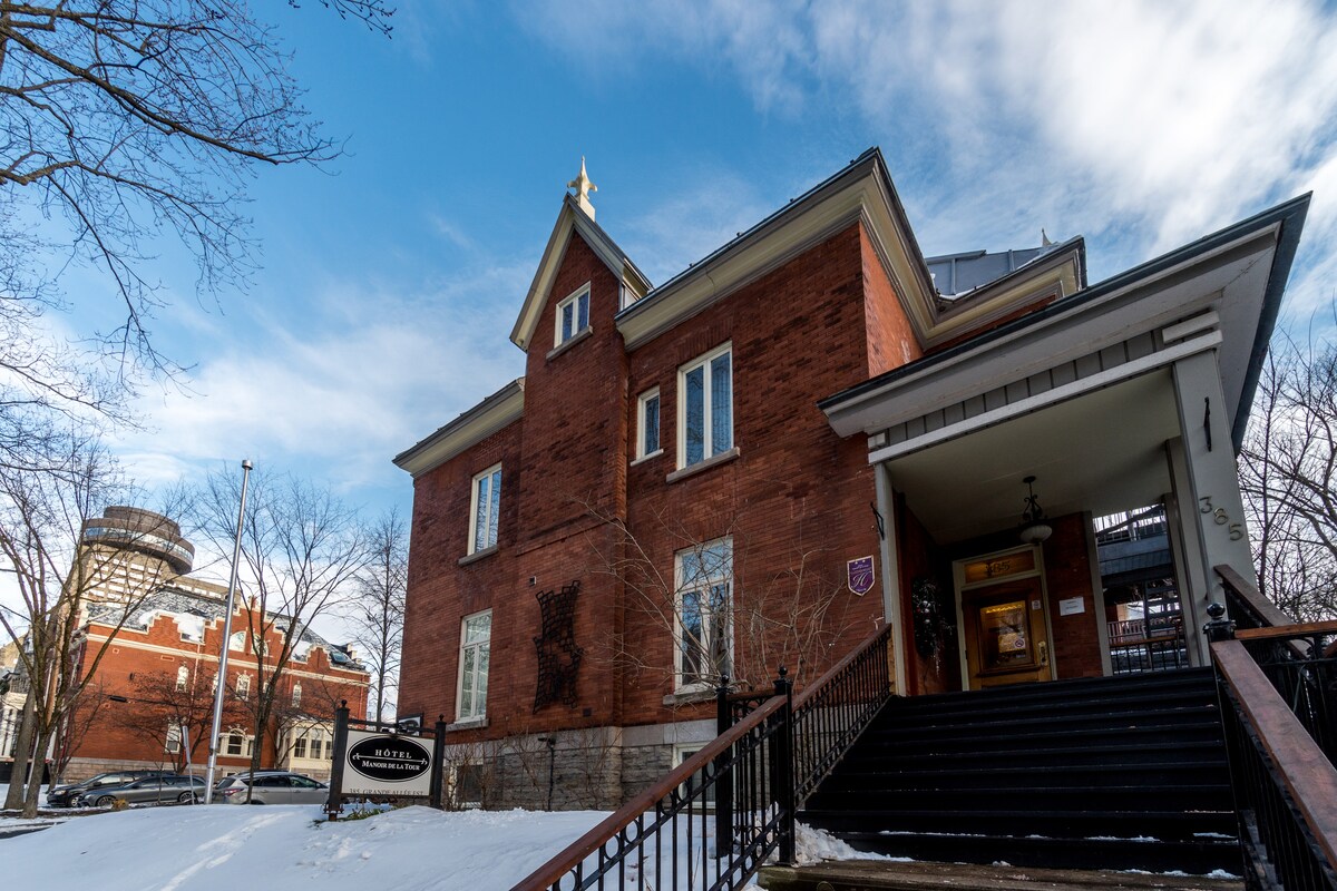 The exterior of a century-old building features a red brick façade, framed by white accents. A decorative gold element crowns the structure, while steps lead to the entrance. Surrounding trees and a glimpse of nearby architecture can be seen, set against a clear sky.