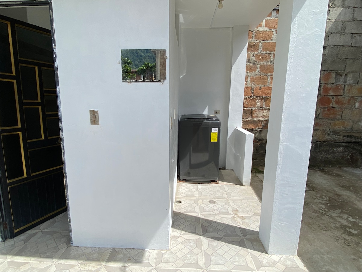 An outdoor utility area is present with a washing machine positioned against a white wall. The flooring features a light-colored geometric tile pattern. The entrance to the space is accessible, with a contrasting black gate visible on the left.