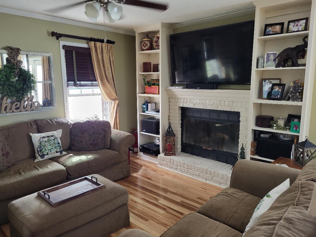 A comfortable living area features a sectional sofa and a coffee table. A large television is mounted above a brick fireplace, surrounded by built-in shelving displaying decorative items. Natural light enters through a window adorned with wood blinds and soft drapes.