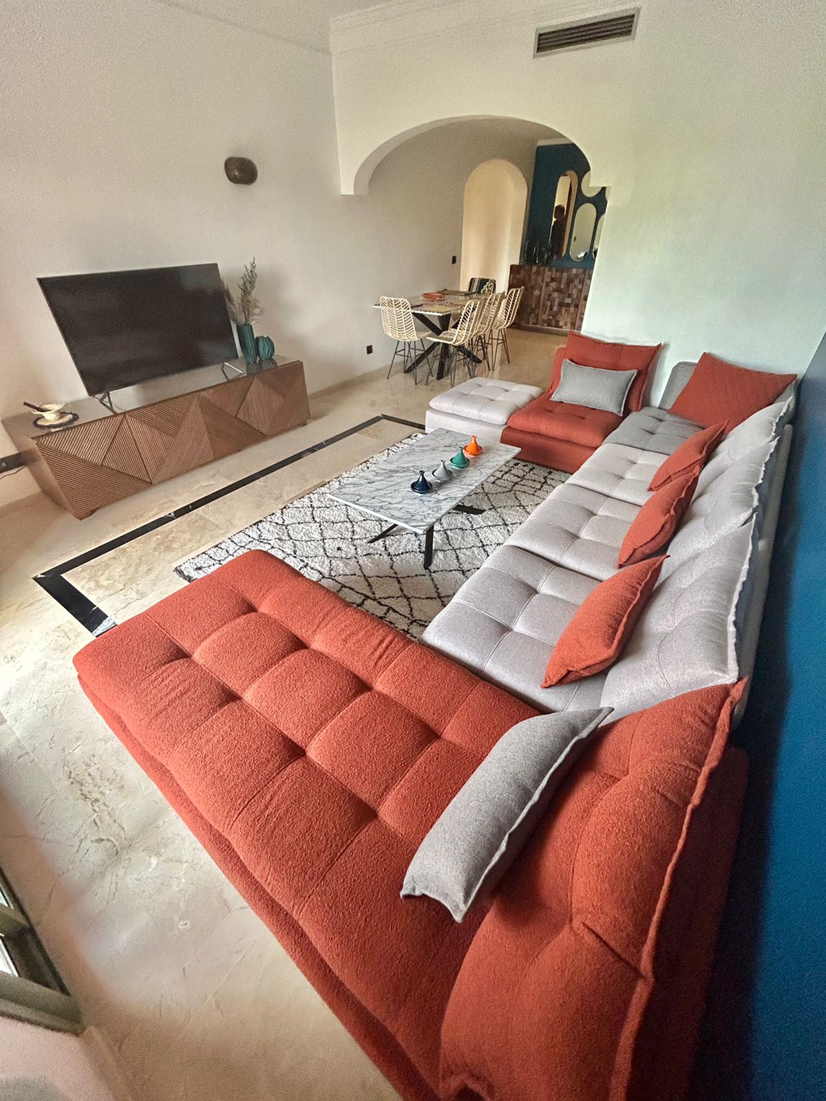 A spacious living area features a large, comfortable sofa with a mix of orange and gray cushions. A flat-screen TV is positioned on a decorative media console. A light-colored rug anchors the seating area, and a dining table is visible in the background.
