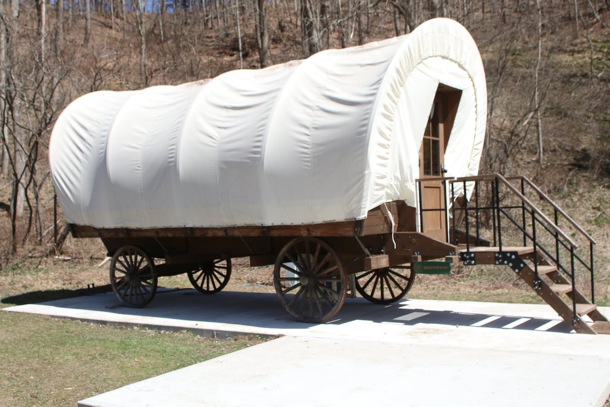 A classic Conestoga wagon is situated on a stone patio, featuring a rounded white canvas cover and wooden framework. A set of metal stairs leads up to the entrance, set against a natural backdrop of trees and hills.