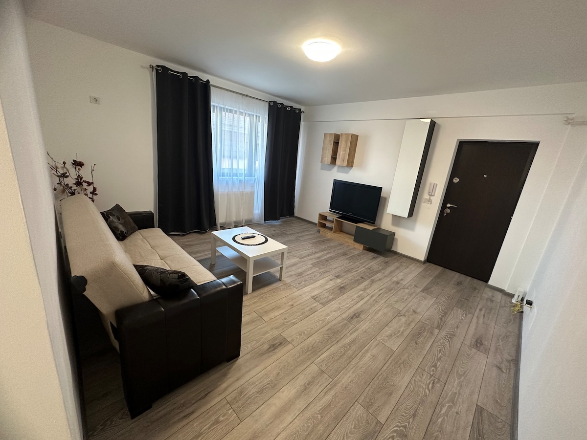 A living area is presented with a light-colored sofa and a small coffee table at the center. A television is positioned on a low media unit against the wall. Natural light enters through a window, framed by dark curtains, enhancing the wooden floor's warm tones.