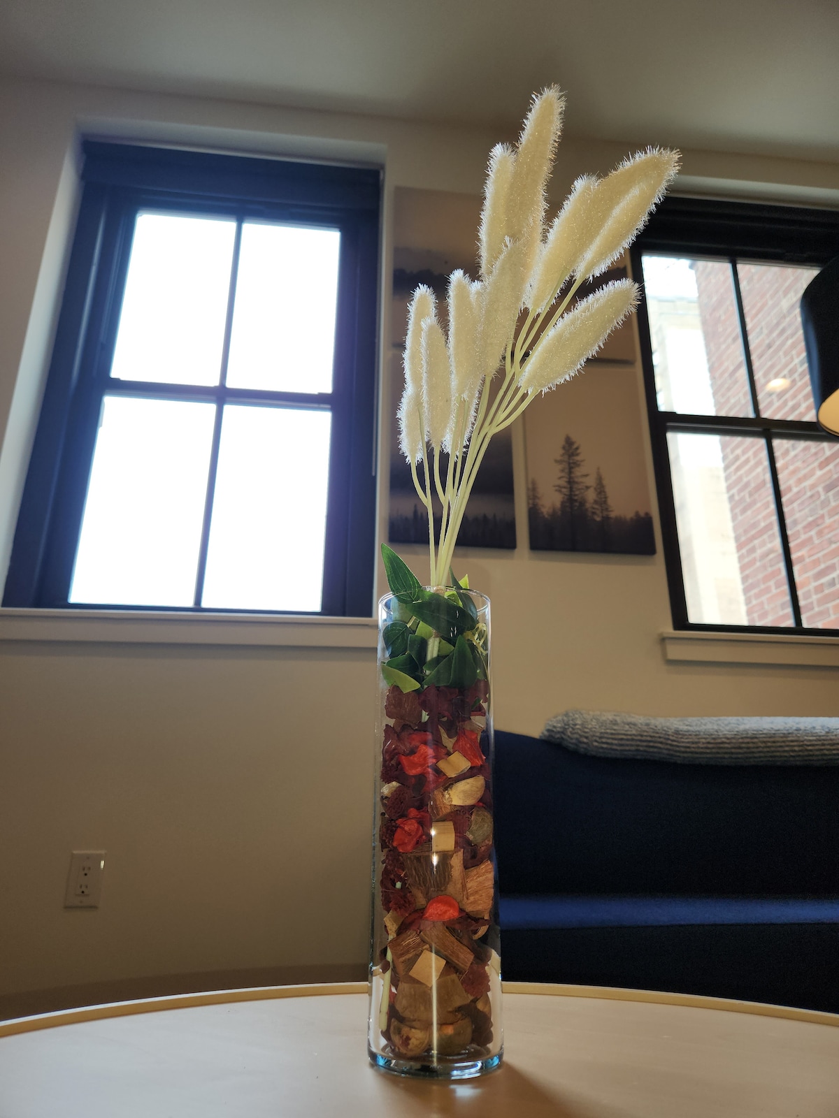 A tall, clear vase holds decorative branches and green foliage, placed on a simple table. Natural light filters through the nearby windows, highlighting the warm tones of the materials and creating a serene focal point within the space.