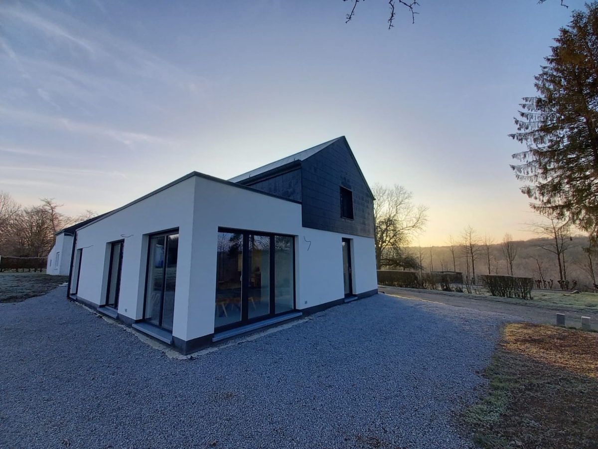 The modern building presents a clean, angular design with large glass windows, allowing for ample natural light. The surrounding gravel area is bordered by greenery, while a tranquil natural setting is visible in the background, showcasing trees against a soft sunset sky.