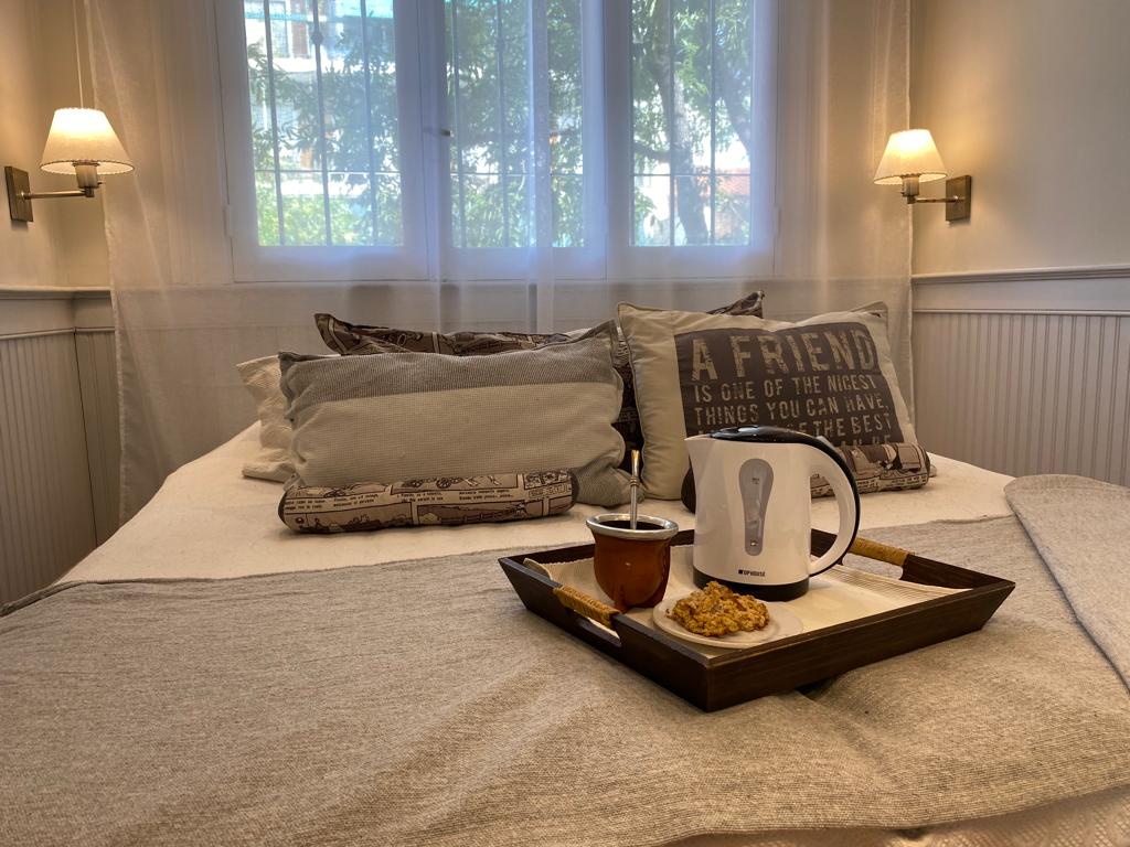 A cozy sleeping area is featured, highlighted by a spacious bed adorned with decorative pillows. A tray holds a teapot and a snack, positioned on soft bedding. Natural light filters through a window, enhancing the serene atmosphere created by the gentle lighting on the walls.