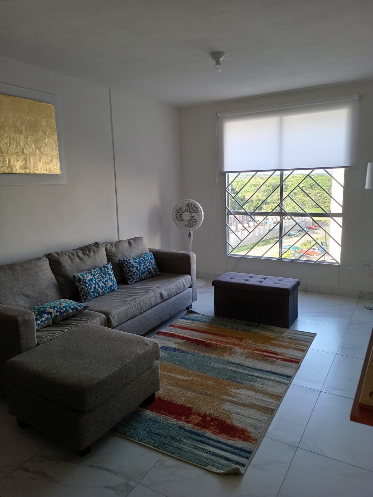 A living area features a gray sofa with colorful throw pillows, accompanied by a small ottoman. A striped area rug adds color to the tiled floor. Natural light enters through a window adorned with a roller shade, providing a view of the outside greenery.