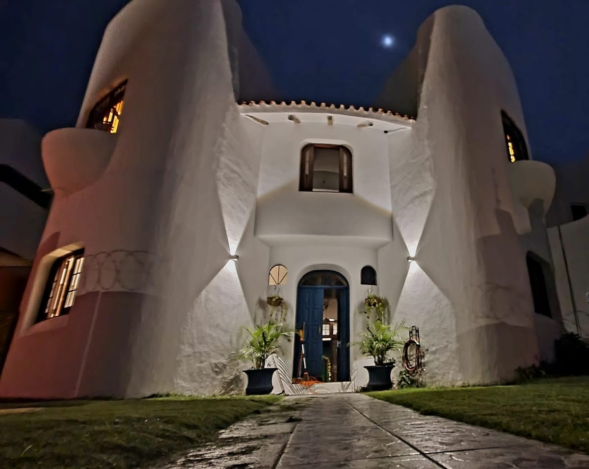 The exterior of the villa is illuminated at night, featuring a white stucco facade and a prominent arched entrance. Decorative plants are positioned beside the doorway, and soft lighting highlights the architectural features while a crescent moon is visible in the night sky.