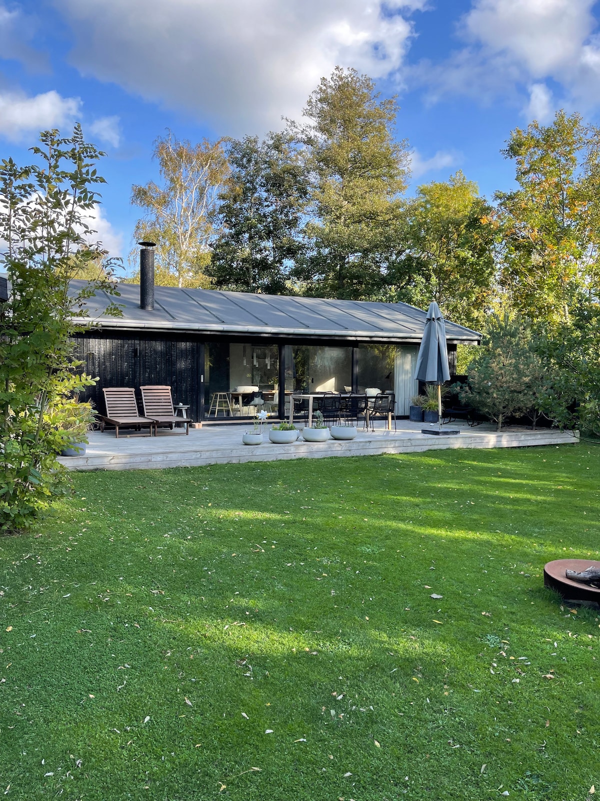 A modern black house features a sloped metal roof and large glass windows. An outdoor patio is furnished with several chairs and a table, accompanied by a large umbrella. Surrounding greenery includes trees and potted plants, with a well-maintained lawn enhancing the outdoor space.