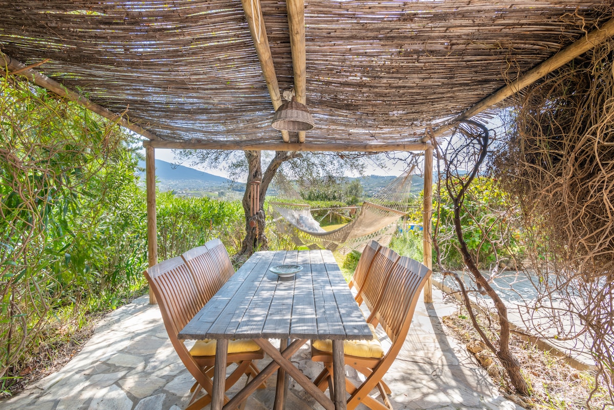 An outdoor dining area is featured beneath a rustic pergola, offering a wooden table surrounded by comfortable seating. Lush greenery frames the space, while a hammock is visible in the background, inviting relaxation in the tranquil surroundings.