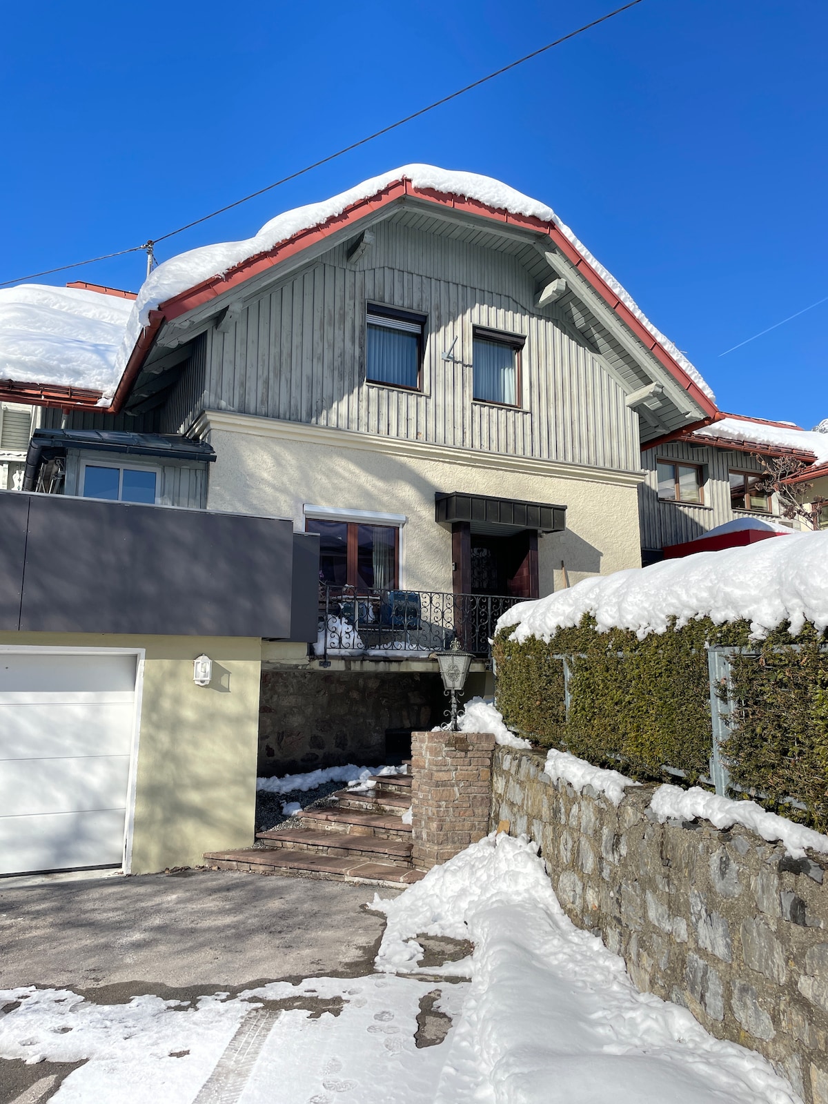 A charming multi-story house is shown, featuring a sloped roof with red trim and snow-covered eaves. Double windows are present on the upper level, while a welcoming entrance is visible at the front. A snow-blanketed driveway leads to the garage, surrounded by manicured hedges.