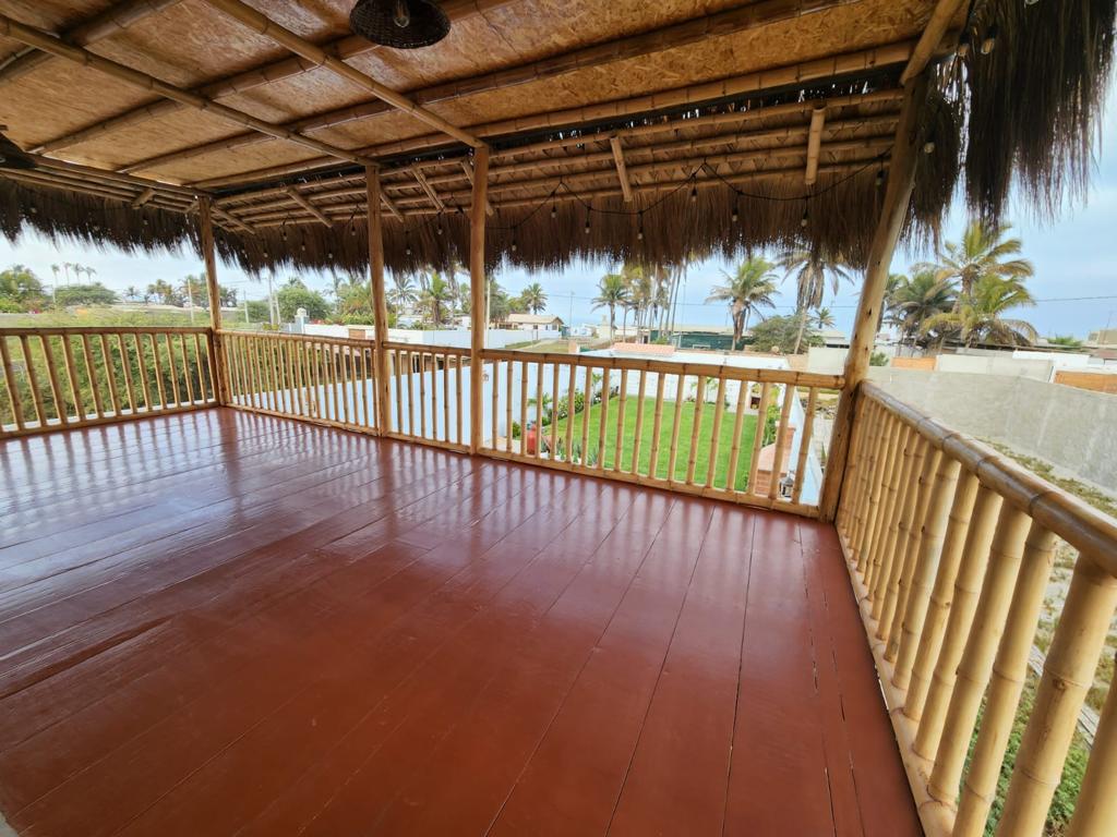 A spacious outdoor deck features bamboo railings and a thatched roof, offering views of the surrounding greenery and ocean. The smooth wooden flooring is well-maintained, creating an inviting area for relaxation. Sunlight filters through palm trees, enhancing the open atmosphere.