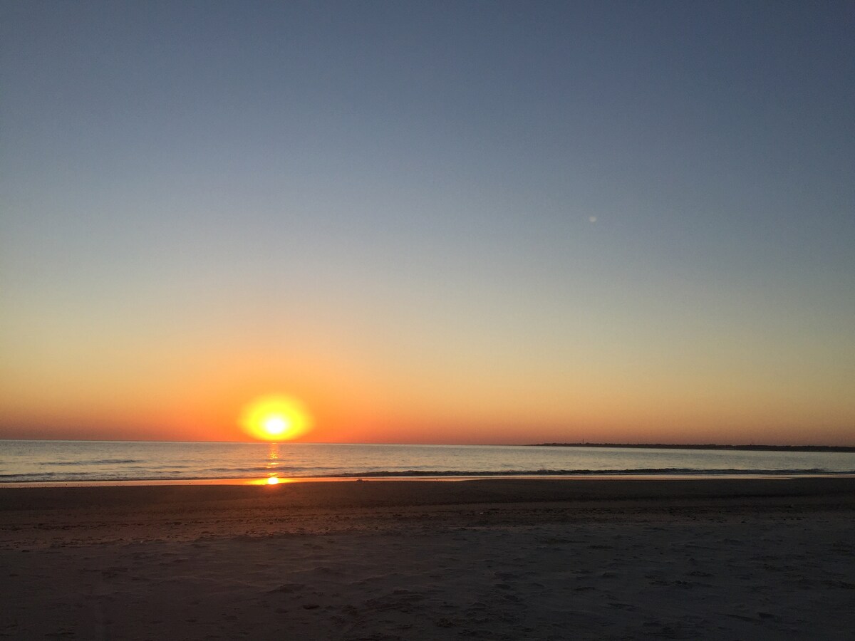 The image captures a serene ocean sunset, with the sun positioned just above the horizon, casting warm orange and yellow hues across the sky. The calm waters reflect the colors, while a smooth sandy beach stretches out in the foreground.