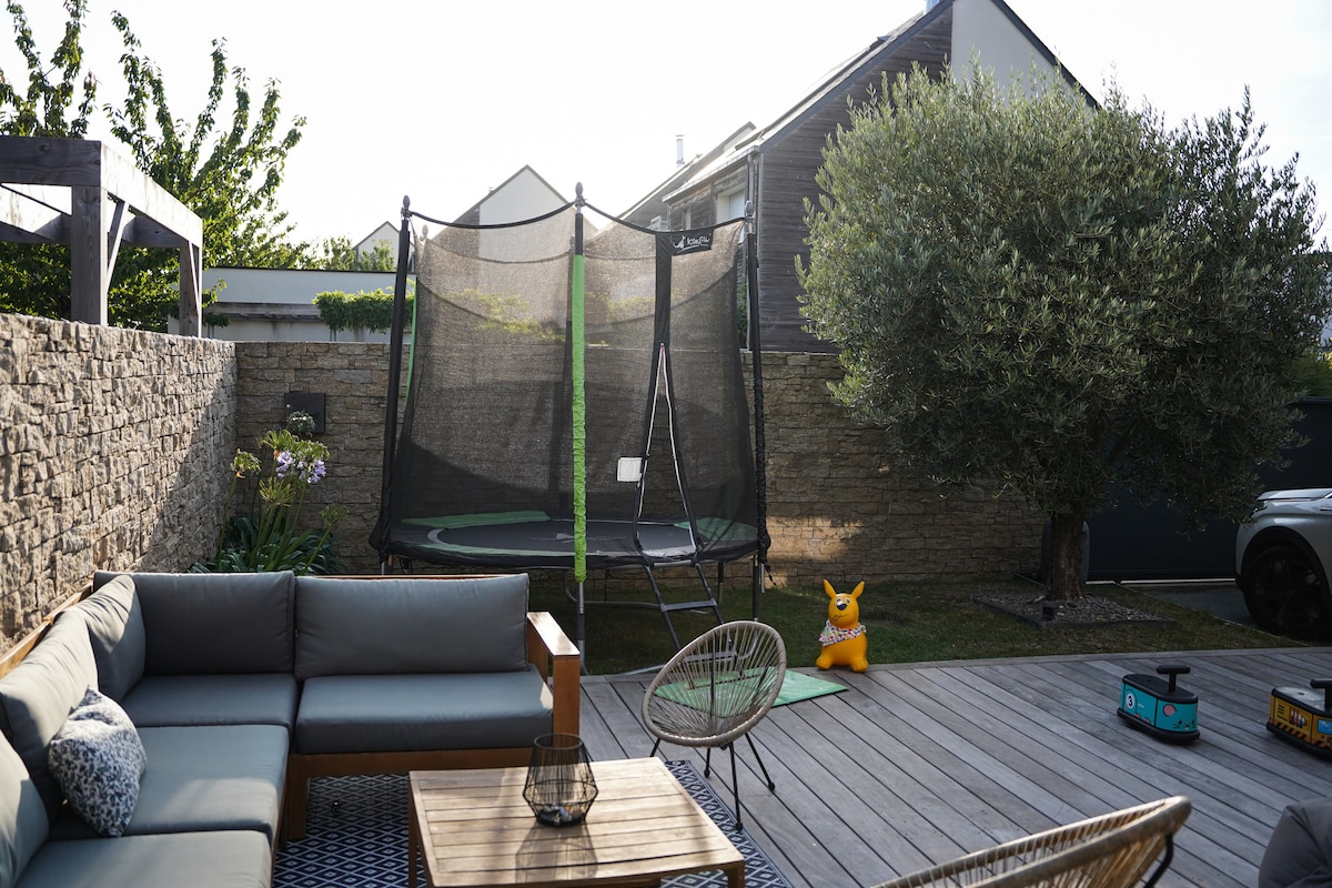 An outdoor seating area is featured, complete with a spacious couch and wooden coffee table. A trampoline is visible in the background, surrounded by greenery. A vibrant children's toy is positioned nearby, complemented by a stone wall that adds texture to the tranquil setting.