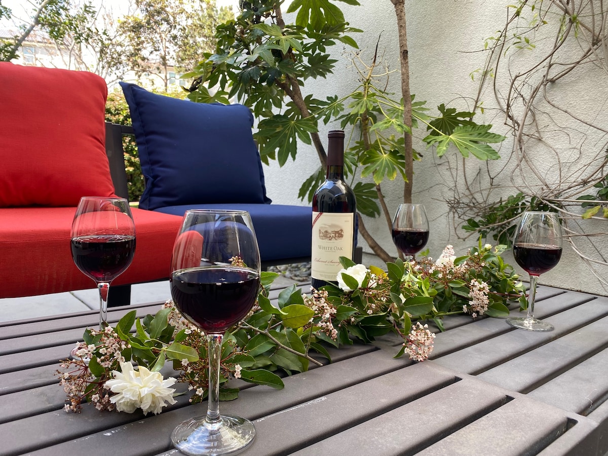 A cozy outdoor seating area features two colorful chairs in red and blue. A wooden table displays a wine bottle surrounded by four glasses filled with red wine. Green foliage and flowers add a touch of nature to the space.