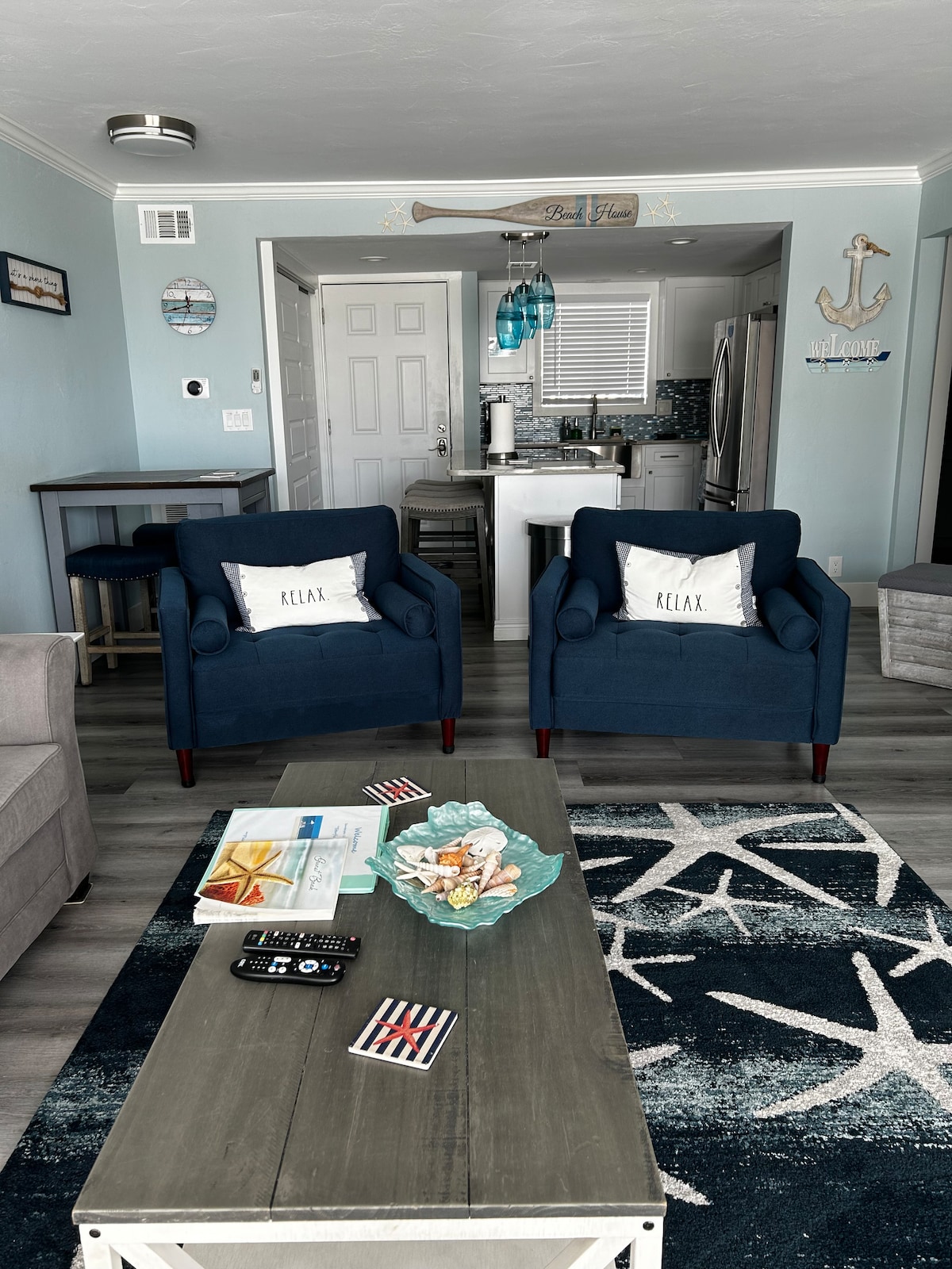 A living area features two blue armchairs with decorative pillows, situated beside a light gray sofa. A coffee table displays seashells in a glass bowl. A patterned area rug enhances the space, while a view into the kitchen reveals a bright and airy layout with coastal decor.