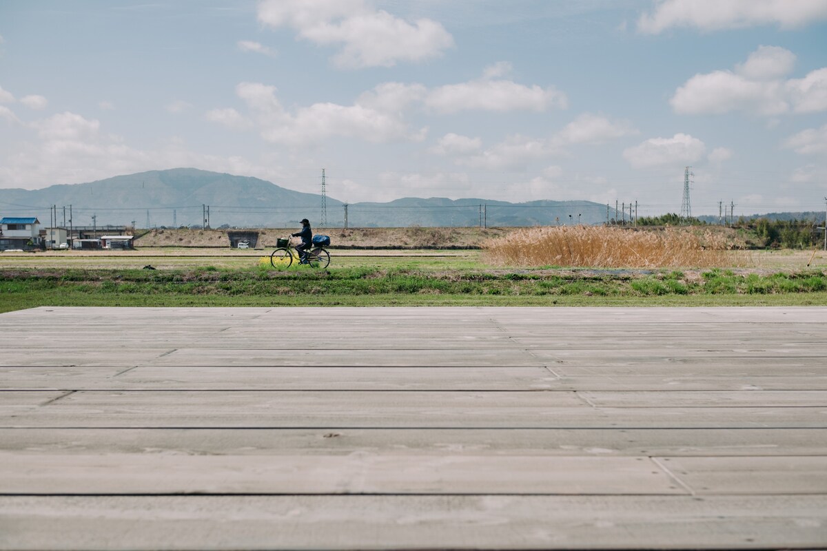 A cyclist rides along a path bordered by lush greenery and golden reeds. In the background, hills are visible under a partly cloudy sky, with distant structures adding to the rural landscape.