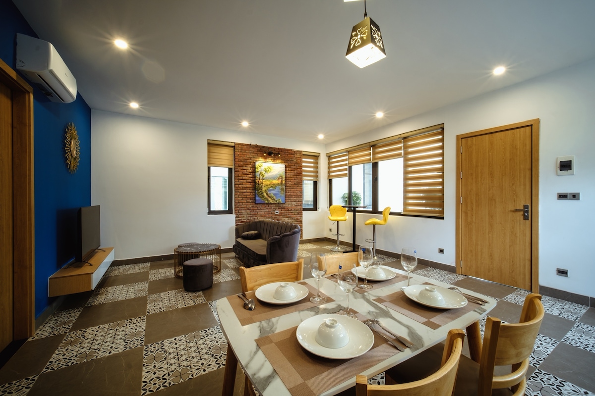 A modern living area features a stylish dining table set with plates and glasses. A comfortable sofa and two bar stools complement the space. Large windows allow natural light to fill the room, while artwork adorns the wall, accentuating the contemporary decor.