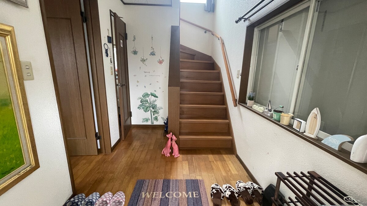 An inviting entrance area features warm wooden flooring and a staircase leading to the second floor. A welcome mat is placed at the base of the stairs, surrounded by various indoor slippers. Natural light fills the space through large windows, enhancing the overall openness.