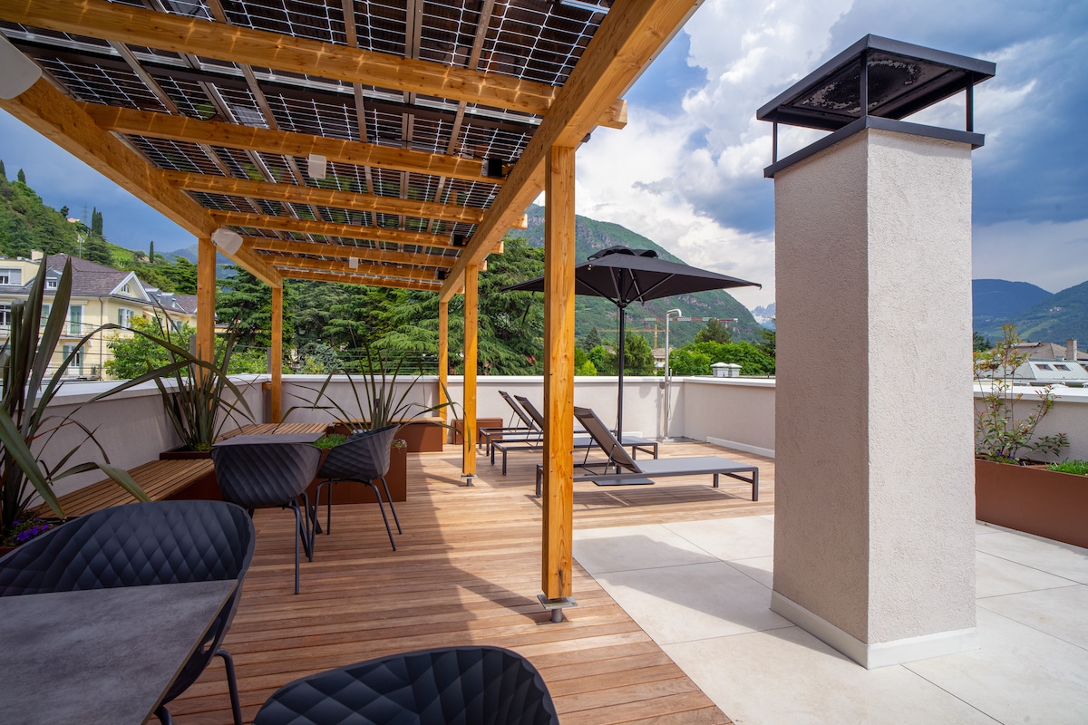 A rooftop area is framed by wooden beams and includes comfortable lounge chairs and a large umbrella. Potted plants line the space, adding greenery to the outdoor seating area, while mountain views are visible in the background beneath a partly cloudy sky.