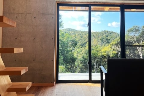 A large glass sliding door offers views of the lush green mountains, allowing natural light to fill the space. A wooden staircase is visible to the left, enhancing the modern design of the interior.