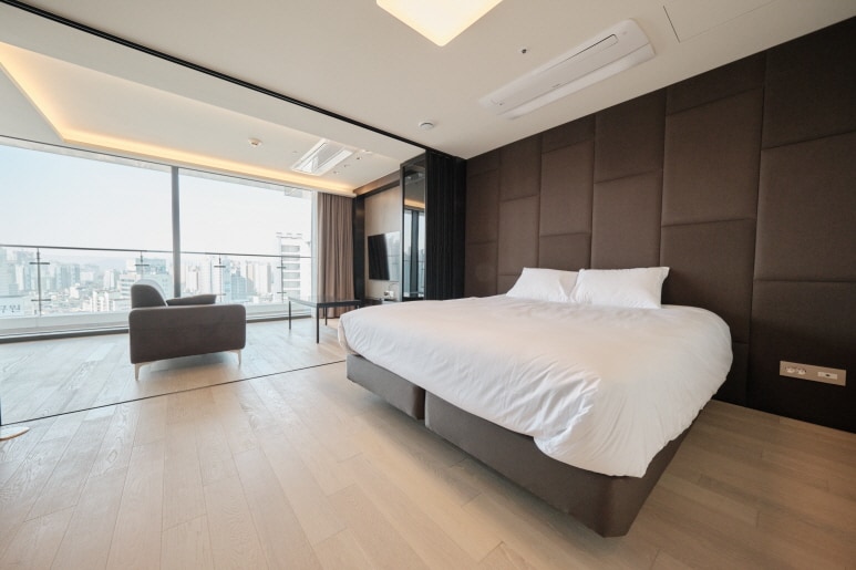 A spacious bedroom featuring a large bed with white bedding set against a dark accent wall. A comfortable chair is positioned nearby, and floor-to-ceiling windows provide a view of the city skyline. Natural light floods the room, highlighting the sleek wooden flooring.