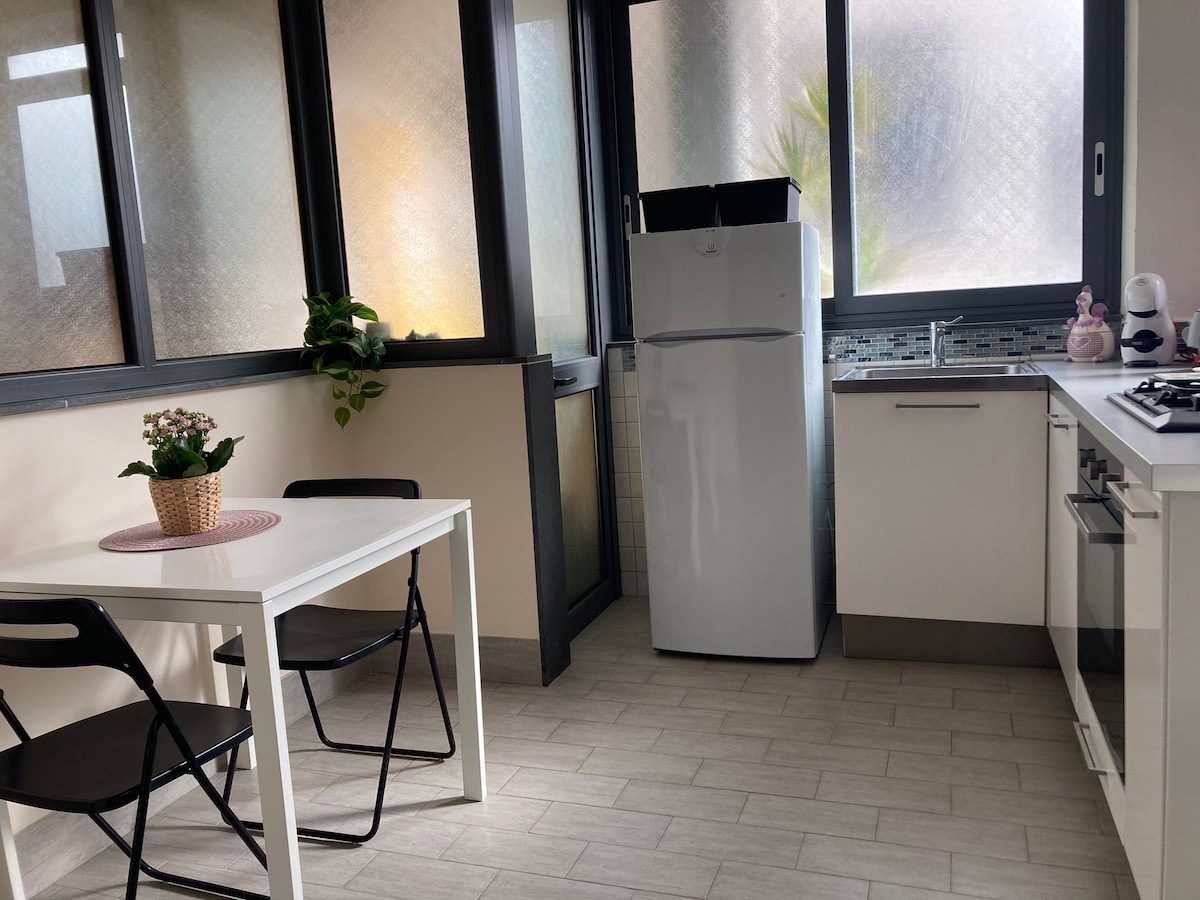 A bright kitchen area is visible, featuring a white dining table with two black chairs. A large window allows natural light to fill the space. The kitchen is equipped with a refrigerator and essential appliances, offering a clean and modern design.