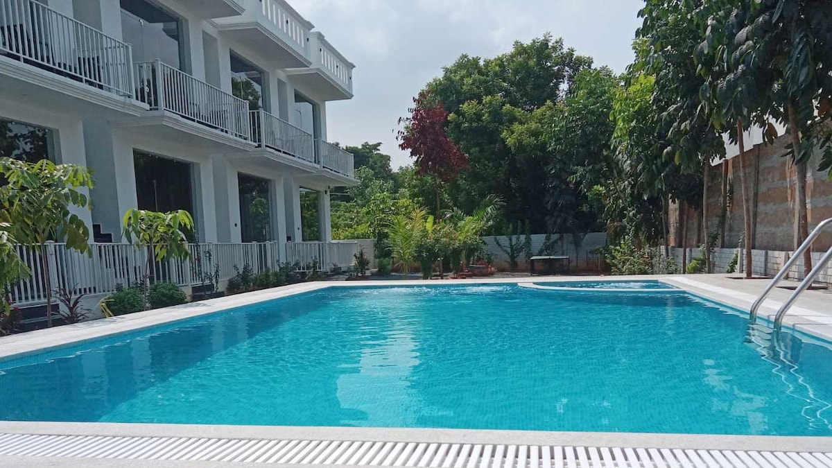 A clear swimming pool occupies the foreground, reflecting the blue sky above. Lush greenery surrounds the area, with vibrant trees and plants enhancing the natural setting. The resort's white building features spacious balconies overlooking the pool, contributing to a serene atmosphere.