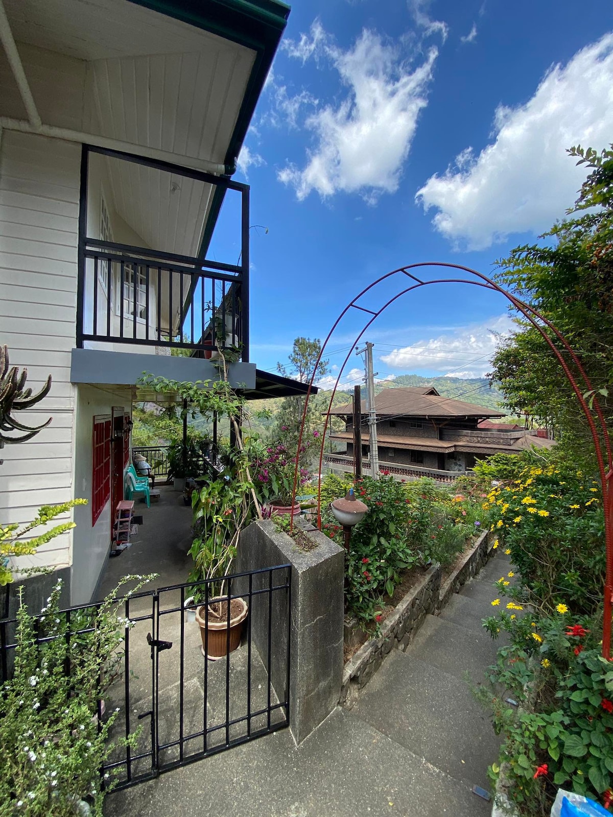 A charming entrance features a decorative archway, leading to a path surrounded by lush greenery and colorful flowers. The exterior of a two-story house is visible, complemented by a balcony that overlooks the tranquil surroundings.