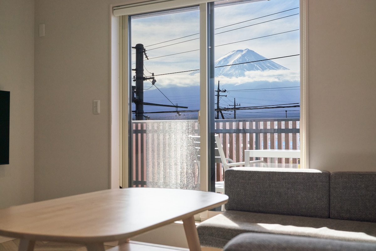A view of Mount Fuji is framed by expansive glass doors, with a transition from indoor to balcony. A light-colored table sits in the foreground, accompanied by a gray sofa, creating a serene space that connects with nature.