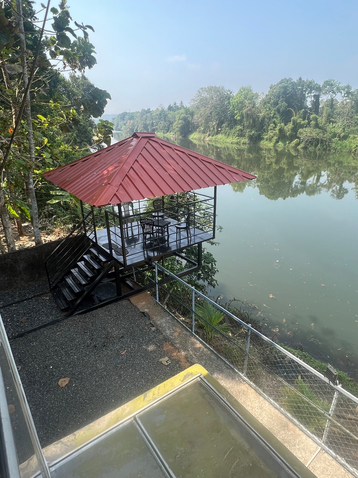 A riverside gazebo with a red roof offers seating for relaxation, overlooking calm waters surrounded by lush greenery. A staircase leads down to the riverbank, enhancing the connection to the natural surroundings.