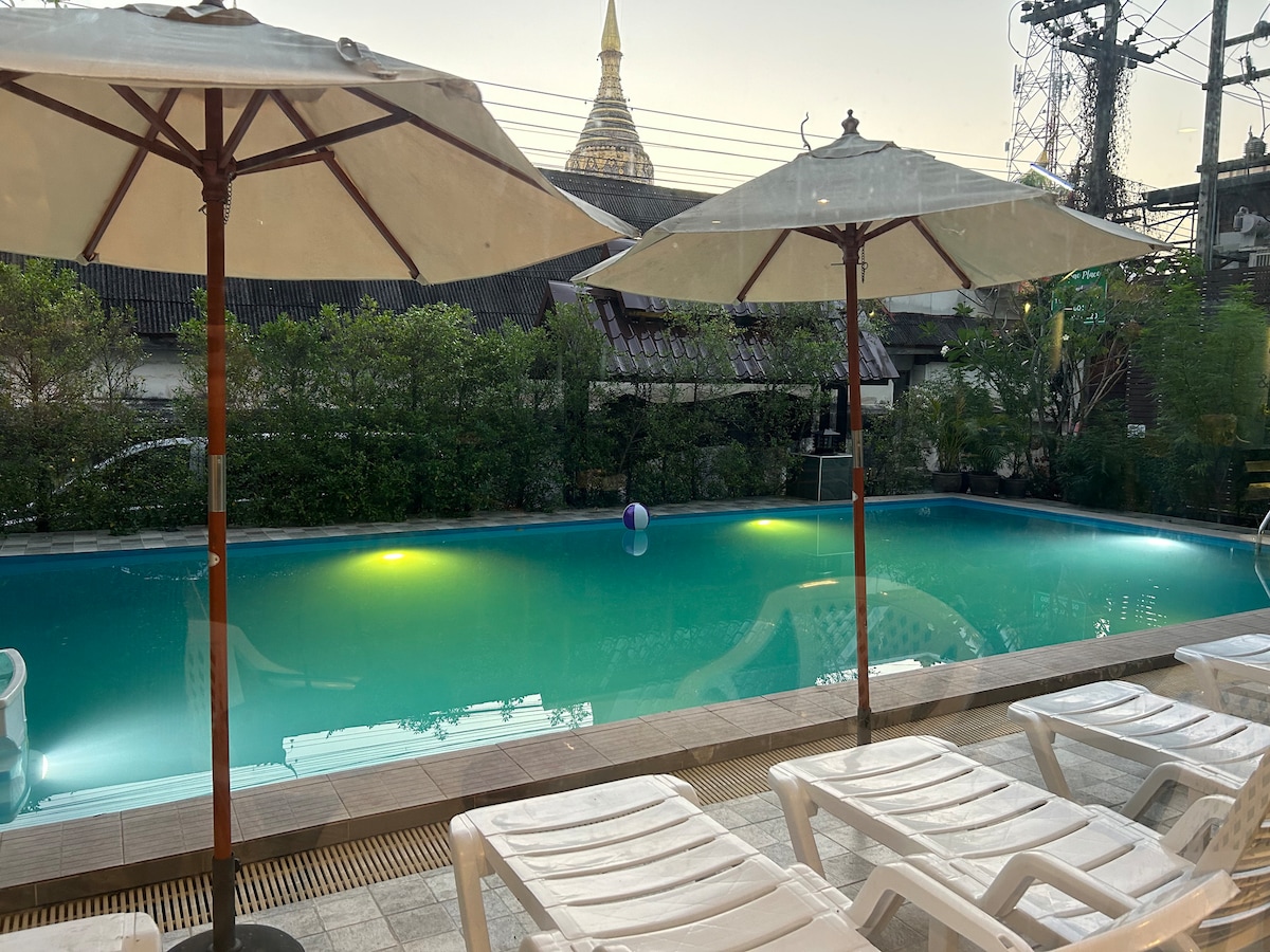 A tranquil swimming pool is framed by two large umbrellas, with lounge chairs arranged nearby. The water's surface reflects the surrounding greenery and evening light, while a soft glow from underwater lights adds to the serene atmosphere. A colorful beach ball floats gently in the pool.