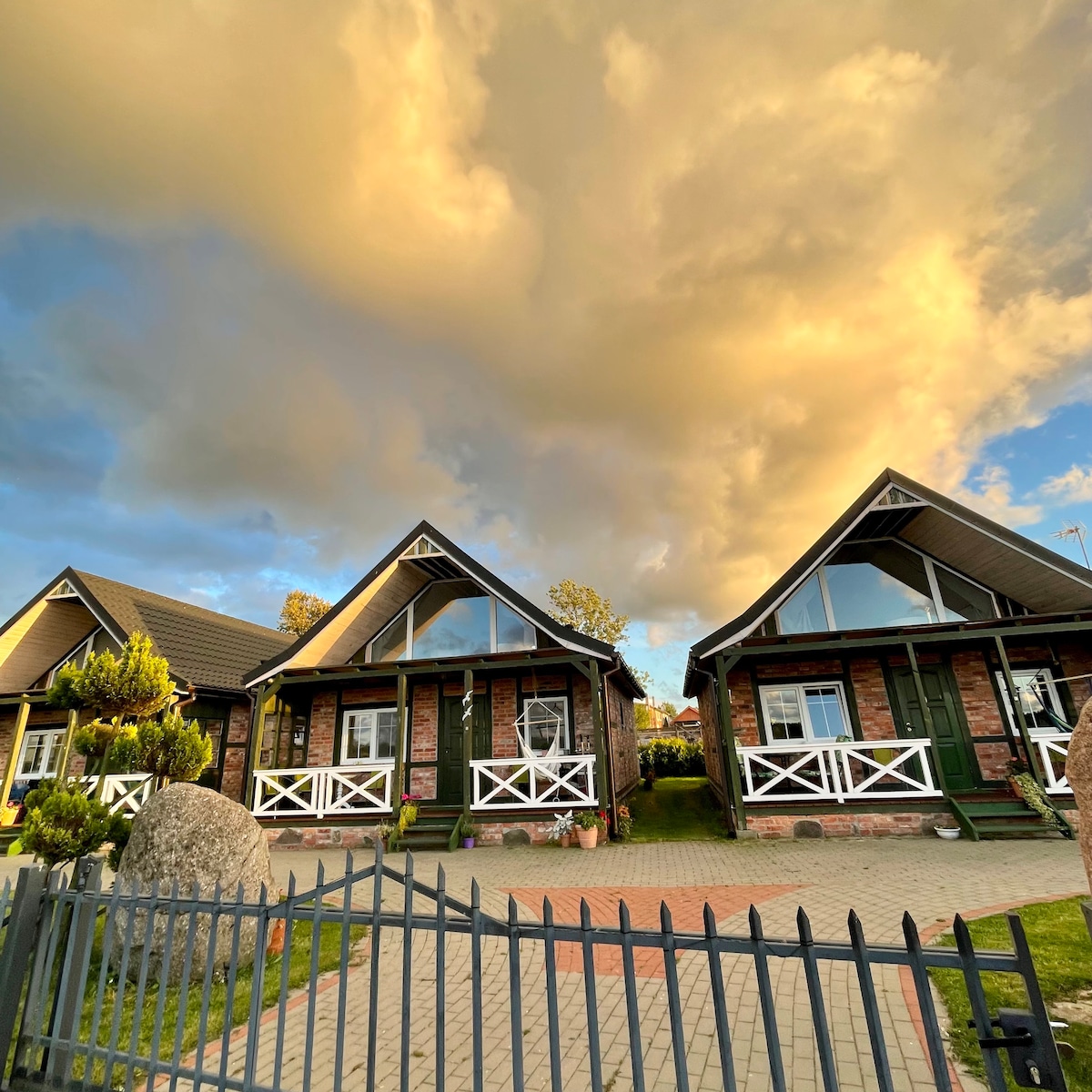 Two charming cottages with sloped roofs are depicted, each featuring large windows and inviting porches. Lush greenery surrounds the properties, with a prominent boulder positioned in the yard. The sky is filled with dramatic clouds, adding depth to the serene setting.