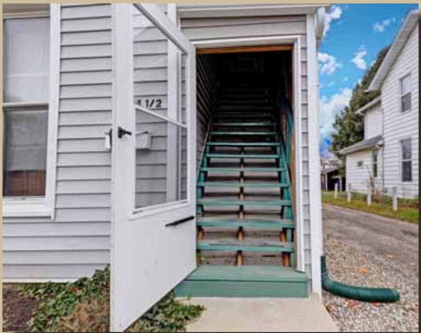 An entrance features a white door that opens to a staircase leading up, surrounded by light gray siding. Natural light filters through nearby windows, creating a welcoming connection between the indoor stairs and the external pathway.
