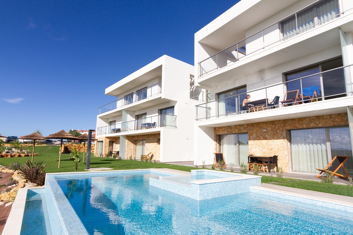 The sleek white building features multiple balconies with seating, overlooking a clear blue saltwater pool. The pool area is surrounded by manicured grass and includes shade umbrellas. Lush gardens with tropical plants are visible, enhancing the serene outdoor setting.