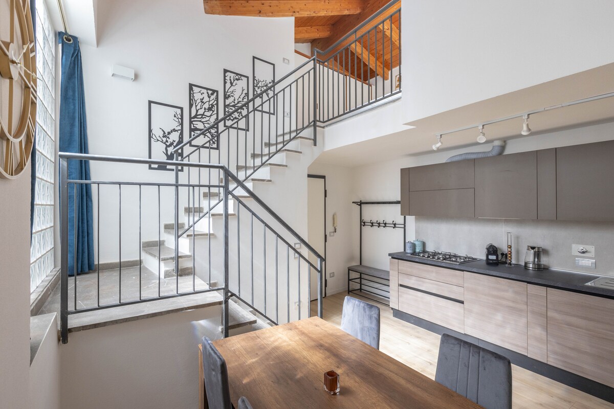 An open-plan living space features a wooden dining table and chairs, with a kitchen area equipped with modern appliances. A staircase leads to the upper level, adorned with artwork against a white wall, while natural light floods in through large windows.