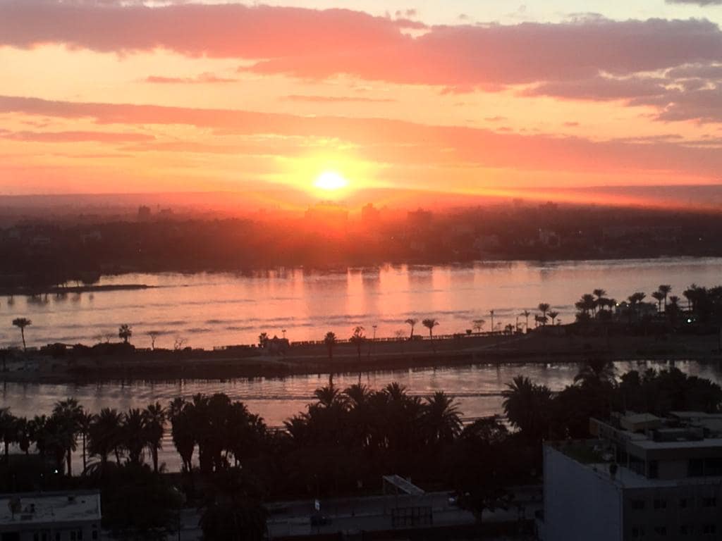 A breathtaking view captures the sunset over the Nile River, displaying shades of orange and pink in the sky. Palm trees line the bank, and the tranquil water reflects the vibrant colors, creating a serene atmosphere. The distant city skyline is subtly outlined against the horizon.