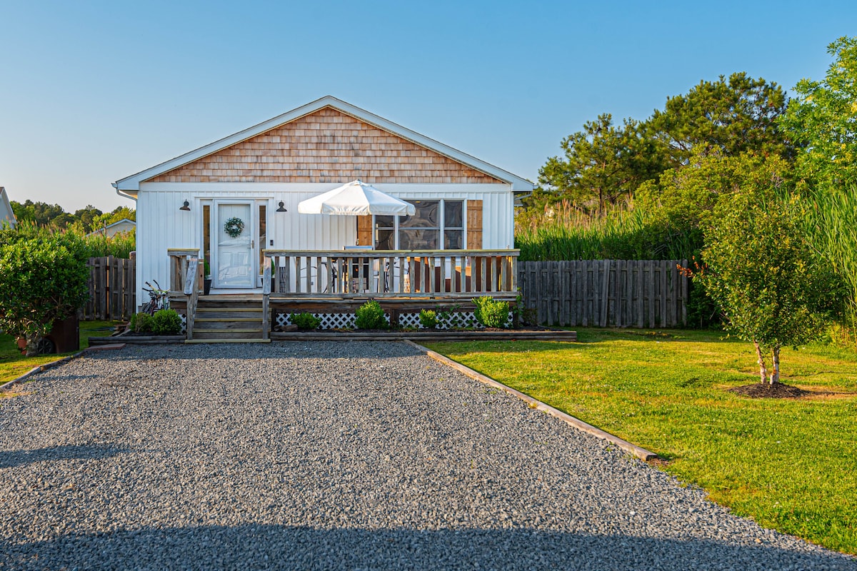 The front exterior of The Cedar Cottage is showcased, featuring a gravel driveway leading to the entrance. A wooden deck is adorned with an umbrella and patio table, complemented by greenery and a wooden fence in the background.