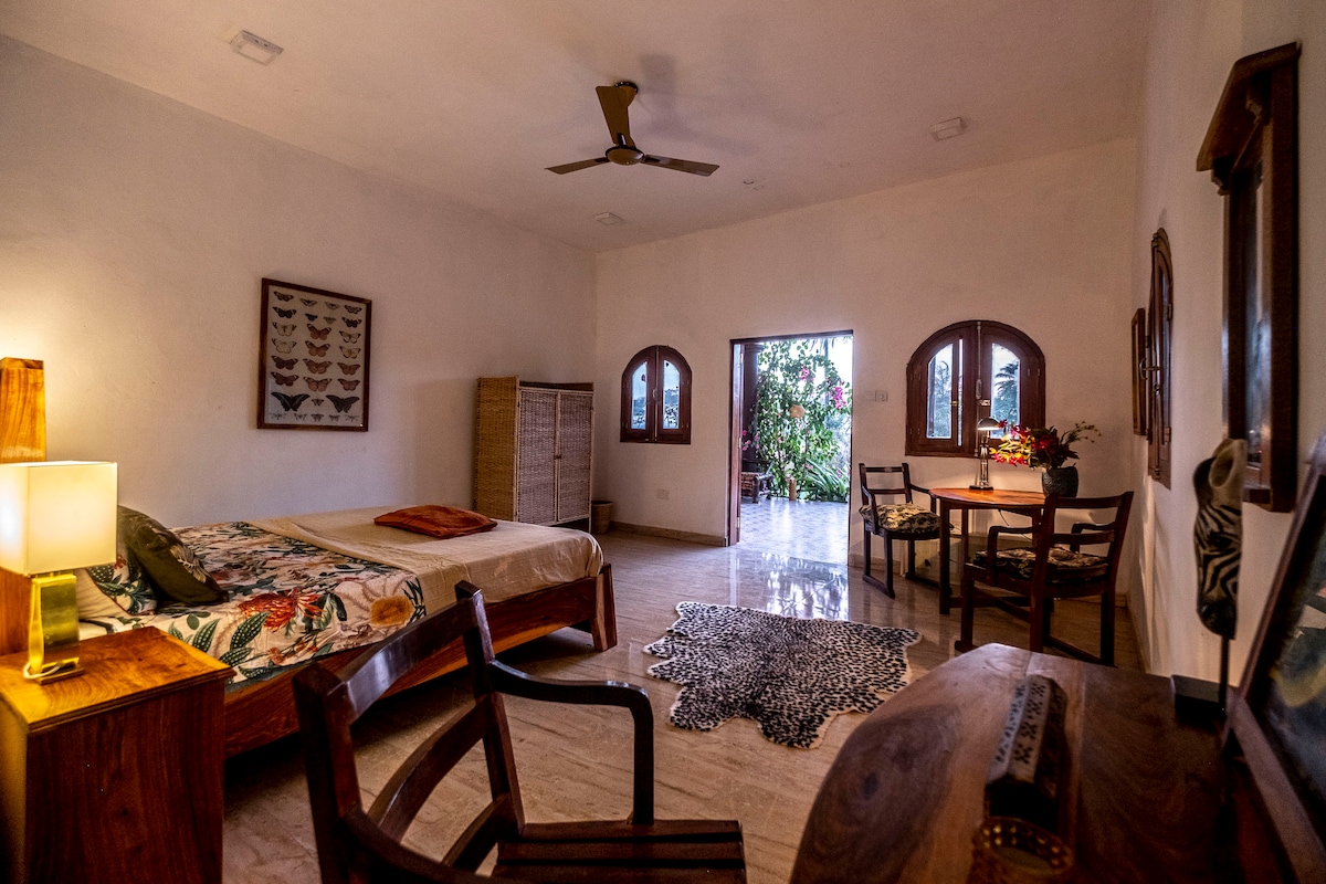 The spacious studio features a queen bed adorned with tropical patterns, complemented by a wooden chair and desk. Natural light enters through arched doorways leading to a lush outdoor area, creating an inviting atmosphere for relaxation.