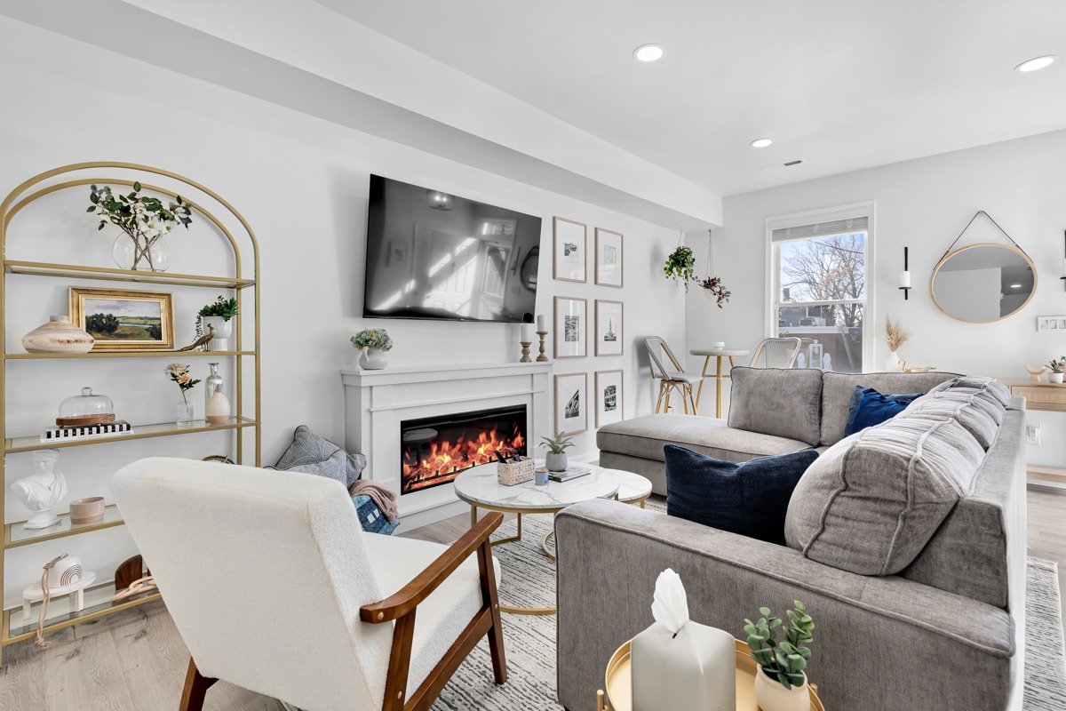 A welcoming living room features a modern grey sectional sofa with navy and white cushions, arranged around a round coffee table. A flat-screen television is mounted above a fireplace, which is framed by white cabinetry. A light-filled window complements the decor, showcasing contemporary artwork.