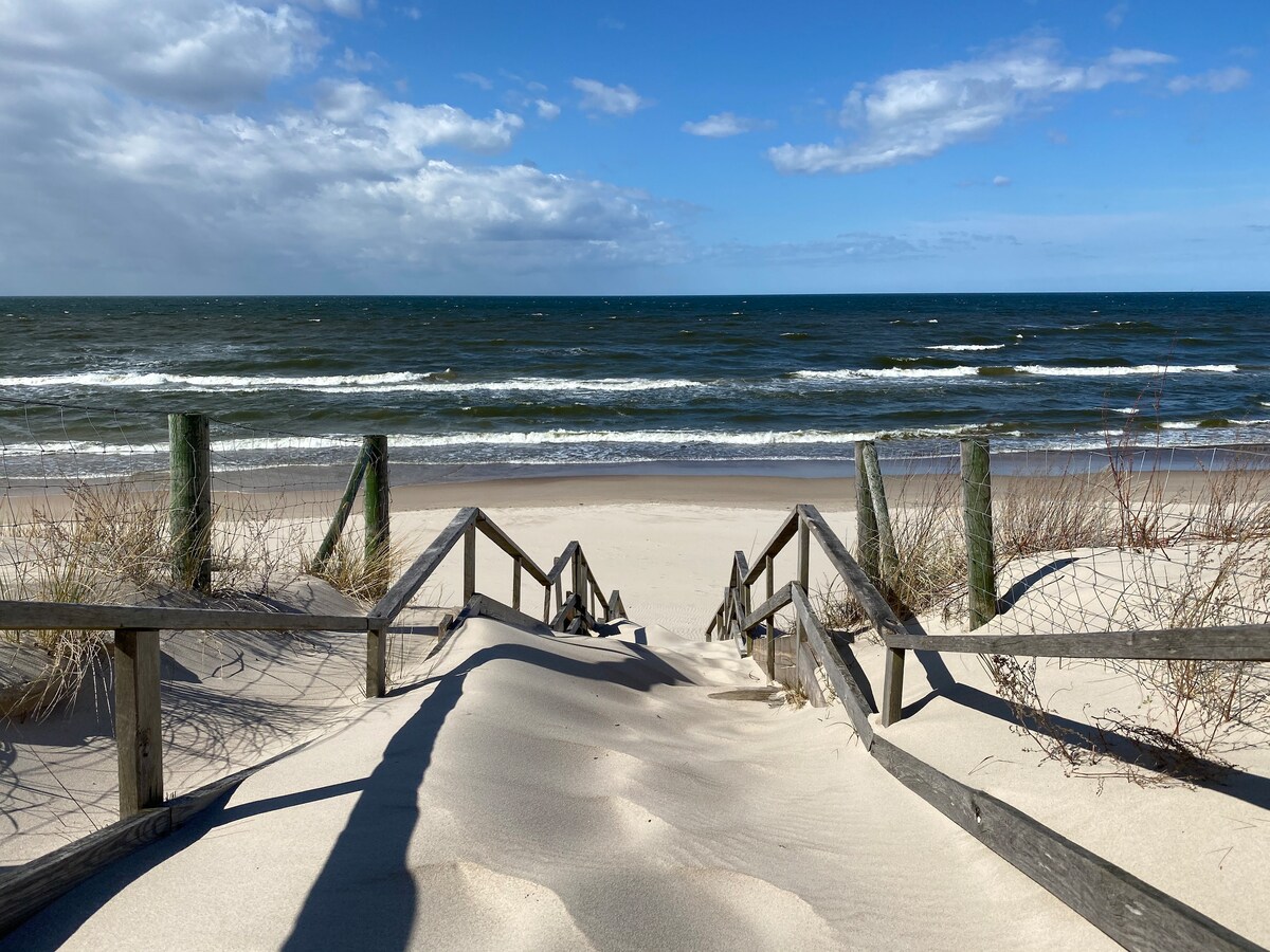 Wooden steps lead down through soft sand to the shoreline, where gentle waves lap at a sandy beach. Tall grasses frame the scene, and a clear blue sky with scattered clouds adds to the coastal ambiance.