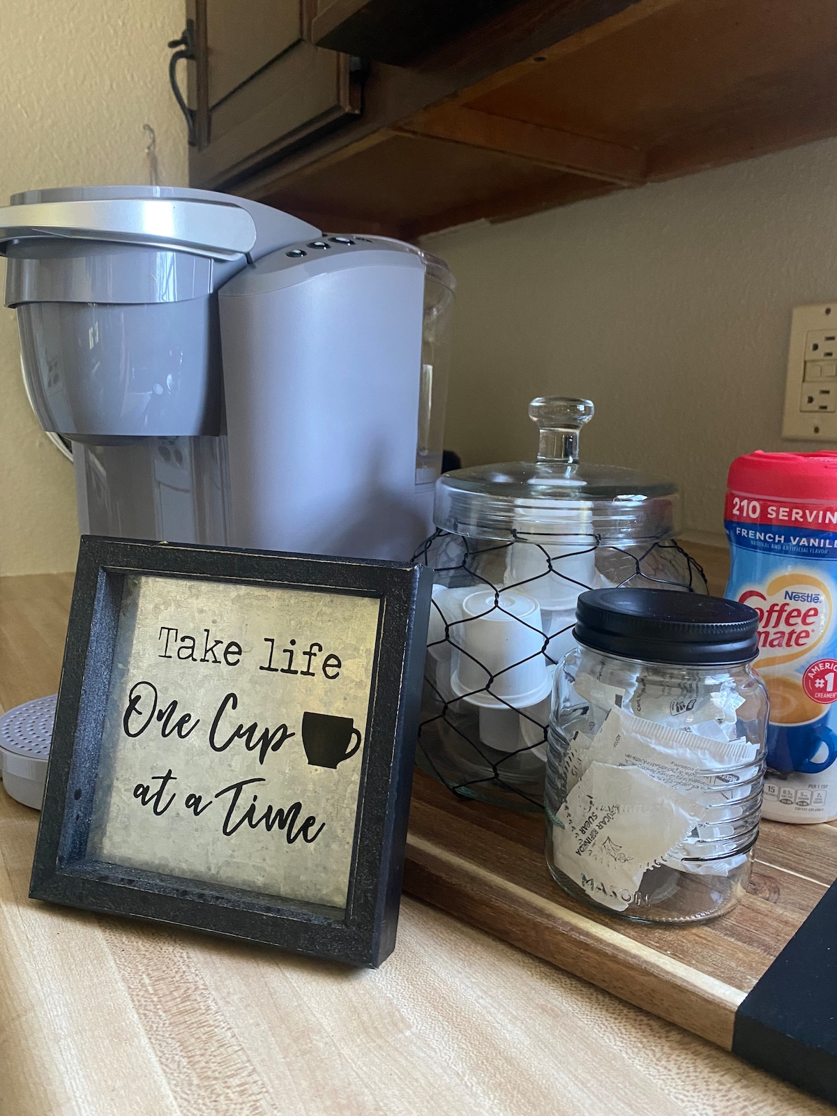 A coffee station is displayed with a single-serve coffee maker and an assortment of creamers. A decorative sign reads 'Take life One Cup at a Time.' Clear containers filled with sugar packets and a jar with tea bags are visible, all arranged on a wooden countertop.