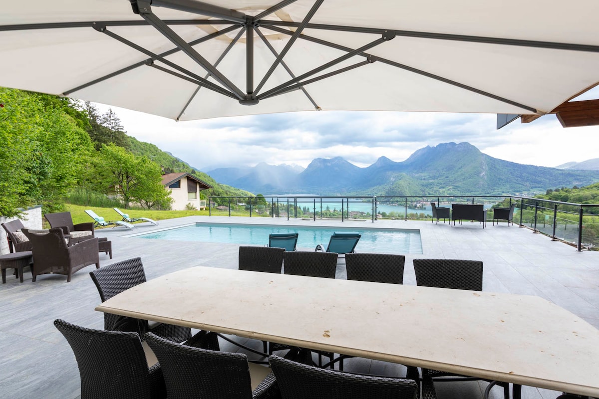 An outdoor dining area features a large table surrounded by chairs, providing ample space for meals. A shaded area is created by a large umbrella, while the pool and scenic mountain views form a beautiful backdrop, enhancing the tranquil outdoor space.
